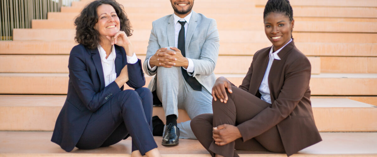Happy business team posing for camera near office building outside. Cheerful multiethnic man and women sitting on stairs and smiling. Successful team portrait concept
