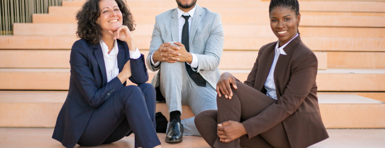 Happy business team posing for camera near office building outside. Cheerful multiethnic man and women sitting on stairs and smiling. Successful team portrait concept